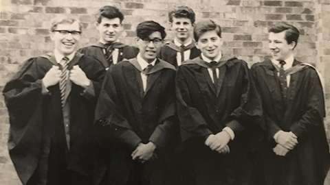 John McGrail stands with a group of five friends. They wear gowns and are standing in front of a brick wall - which is Faraday Hall. 